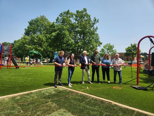 A ribbon-cutting ceremony at the new Plattsville Park Playground. Seven individuals, including Oxford MPP Ernie Hardeman, members of Township Council, and staff, hold a red ribbon across a section of artificial turf in front of new accessible playground equipment. Families and children play in the background under a clear blue sky.
