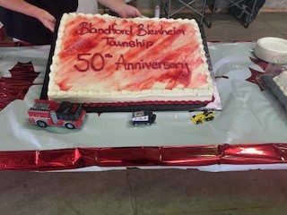 Decorative sheet cake with red and white marbled icing and text reading 'Blandford-Blenheim Township 50th Anniversary.' The cake is on a table with a Canada Day-themed tablecloth, and toy vehicles (fire truck, police car, and yellow car) are placed in front of it.